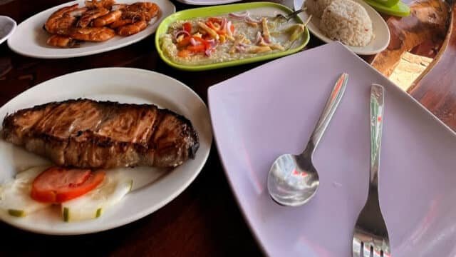 Grilled steak, shrimp, ceviche, and rice served Siargao-style on a wooden table with empty plates and utensils.