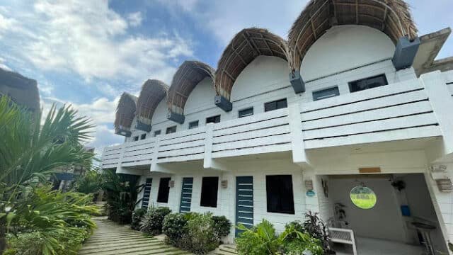 A white two-story building with thatched roof details in Siargao, surrounded by greenery and a cloudy blue sky above.