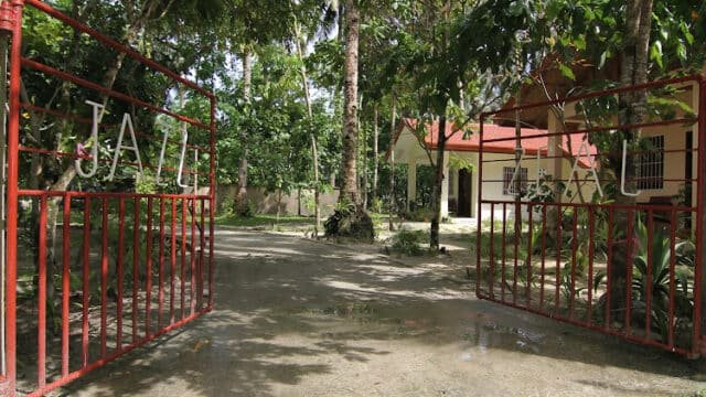 Open red metal gates lead to a house with a red roof in Siargao, surrounded by trees and greenery on a sunny day.