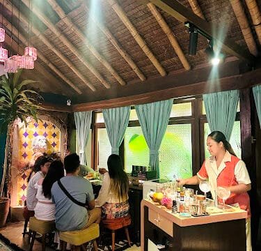 A waitress serves drinks at a cozy, Siargao-inspired tropical cafe where four people sit together at a counter.