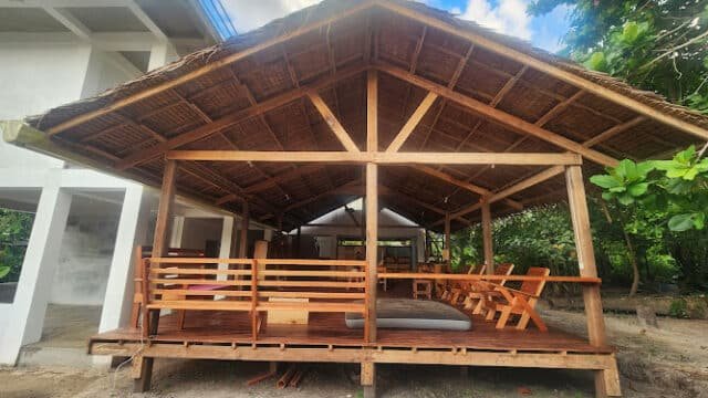 Open-air wooden pavilion with a thatched roof in Siargao, featuring benches, chairs, and surrounded by lush trees and greenery.