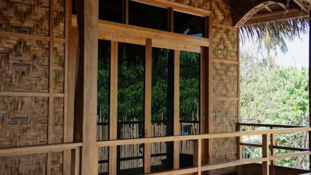 A wooden balcony with woven bamboo walls and a sliding glass door, overlooking Siargao’s lush green trees.