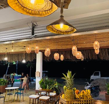 Warm-lit hanging lamps illuminate a cozy, tropical Siargao cafe with a fruit basket and outdoor seating at night.
