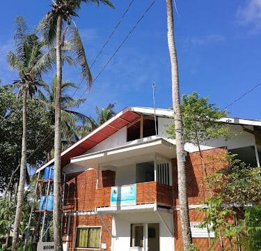 Two-story Siargao guesthouse with bamboo walls, balcony, and palm trees under a bright blue sky.