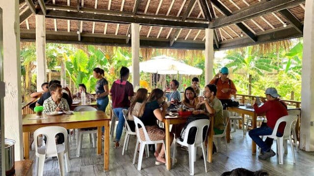 People dining together at wooden tables in an open-air, thatched-roof Siargao restaurant with a dog lounging on the floor.