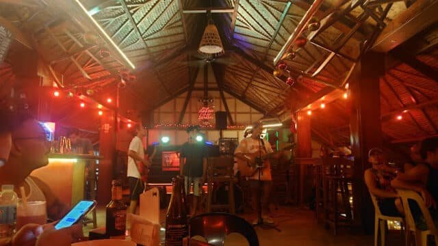 A live band performs on a small stage in a colorful, tropical Siargao bar with people sitting at tables.