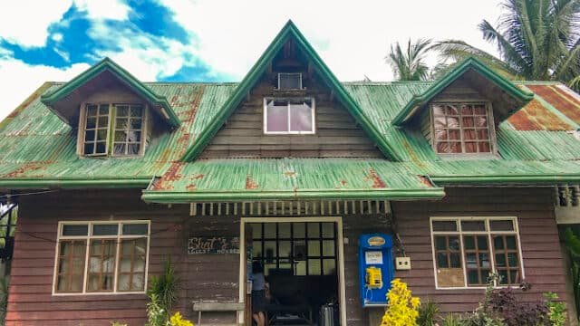 A rustic, wooden house with a green, rusty roof and lush plants in front, reminiscent of Siargao under a bright blue sky.