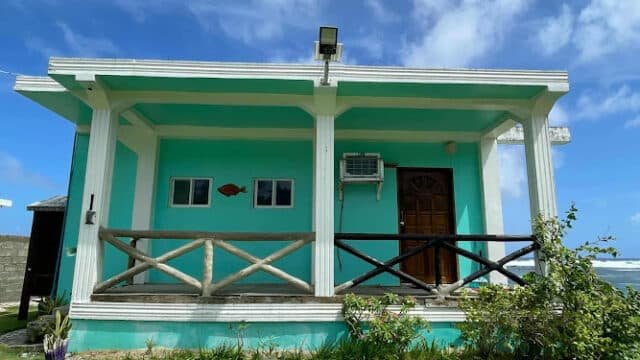Charming small turquoise house in Siargao with a wooden porch railing, door, windows, and an air conditioner under a blue sky.