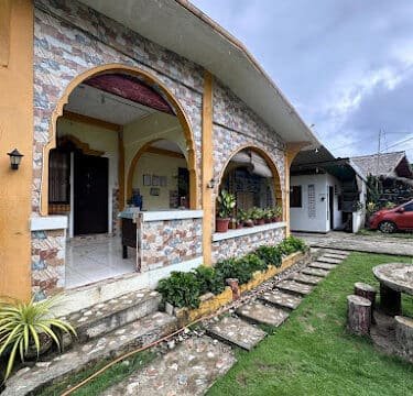 A house with stone archways, a patio, and garden path sits on a grassy yard under Siargao’s cloudy sky.