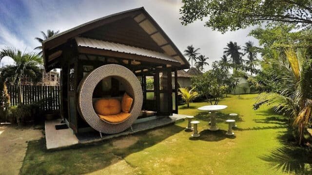 A small wooden gazebo with a round wicker chair in a sunny Siargao garden, surrounded by palm trees and a stone table set.