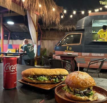 A sandwich, a burger, and a Coke can on a table outdoors in Siargao near a food truck, with string lights overhead.