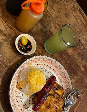 A Siargao-inspired plate with rice, grilled meat, sausage, dipping sauce, condiments, and juice sits on a wooden table.