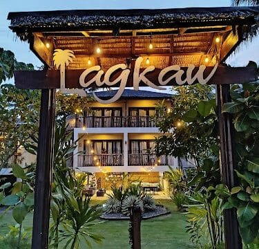 Wooden entrance sign reading Lagkaw with a lit, modern building and lush Siargao greenery in the background at dusk.