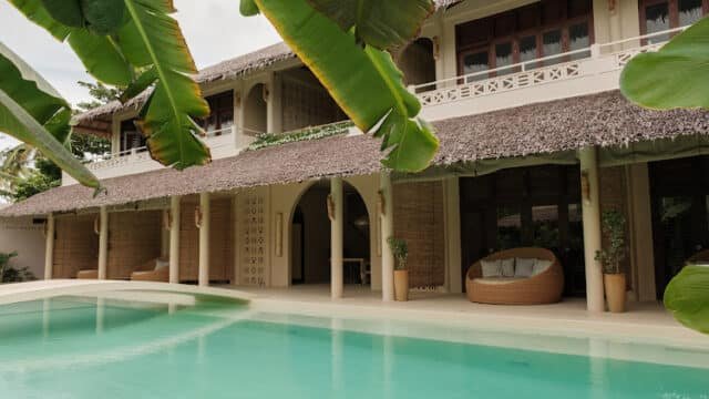 A tropical Siargao villa with a thatched roof, pool, and leafy plants in the foreground.