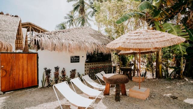 Three white lounge chairs and a thatched umbrella beside a tropical hut amid Siargao’s lush palm trees and greenery.