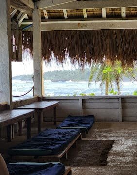 Shaded beachside lounge in Siargao with cushioned benches, a thatched roof, and stunning views of the ocean and palm trees.