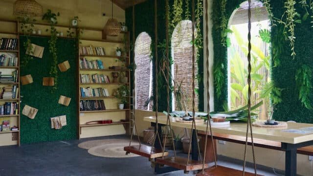 Cozy reading nook with bookshelves, green walls, large windows, and swings as seats around a wooden table—Siargao-inspired charm.