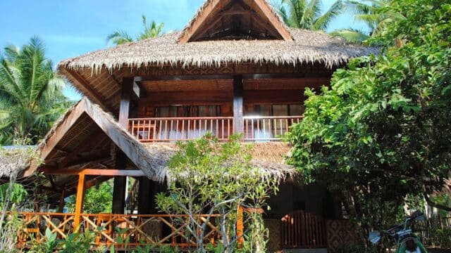 Two-story tropical house in Siargao with a thatched roof, wooden balcony, and lush greenery framing the inviting entrance.
