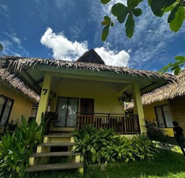 Yellow bungalow with a thatched roof in Siargao, surrounded by greenery under a bright blue sky with clouds.