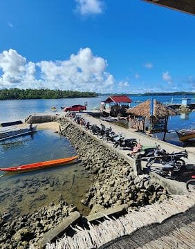 A sunny Siargao dock with boats, motorbikes along a stone pier, and huts by the water under a clear blue sky.