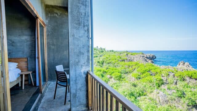A balcony with chairs overlooks Siargao’s green cliffs and blue ocean under a clear sky, next to a modern bedroom.