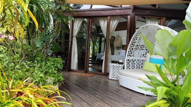 Cozy patio with a white wicker lounge chair, surrounded by lush green plants, exuding tropical Siargao vibes outside a glass-doored room.
