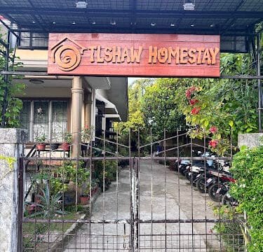 Entrance gate and signboard reading Tshaw Homestay in Siargao, in front of a house with plants and parked vehicles.