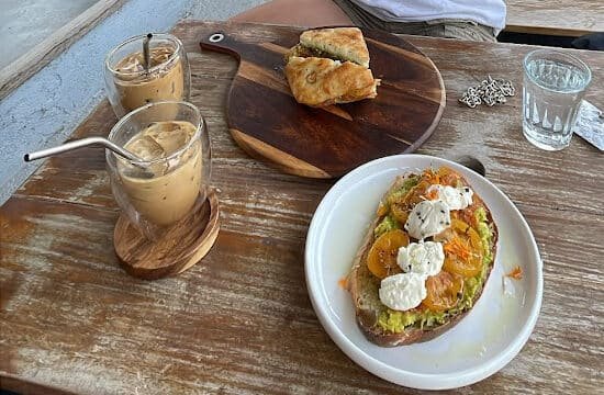 Two iced coffees, Siargao-style focaccia bread, and avocado toast with tomatoes and cream on a wooden table.