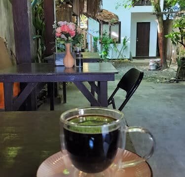 A glass cup of coffee on a saucer sits on an outdoor table in Siargao, surrounded by flowers and lush greenery in the background.