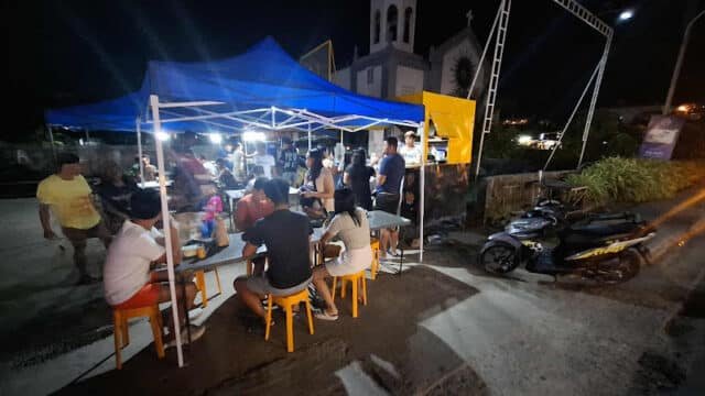 People dining at outdoor food stalls under a blue tent at night in Siargao, with a church visible in the background.
