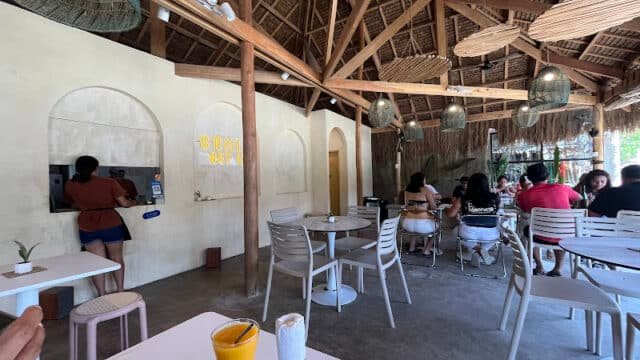 Cafe with wooden ceiling in Siargao, people dining, and a woman using a payment kiosk on a cream-colored wall.