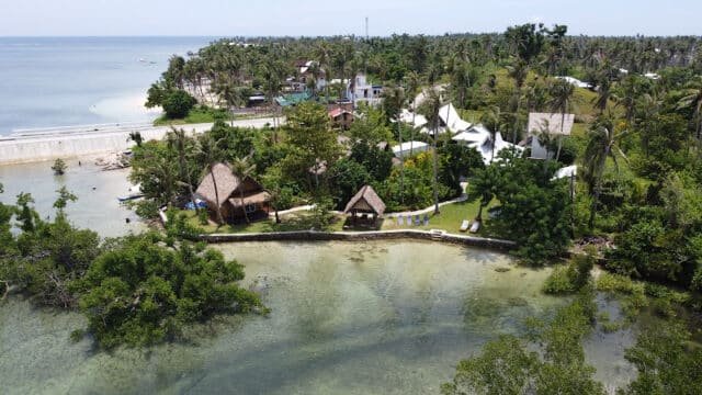 Aerial view of Siargao's coastal village with huts, palm trees, and clear shallow water along the shoreline.