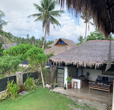 A tropical Siargao backyard with palm trees, a thatched roof patio, and green grass under a cloudy sky.