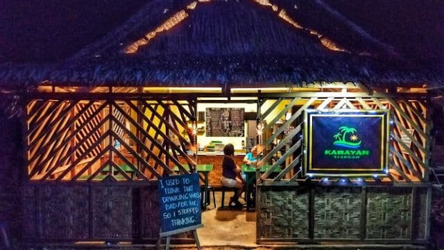 A warmly lit bamboo restaurant in Siargao at night, with two people inside and a humorous sign by the entrance.
