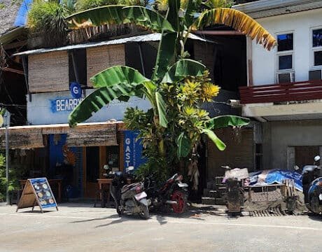 A tropical Siargao street with a guesthouse, banana trees, parked scooters, and a bright blue sky.