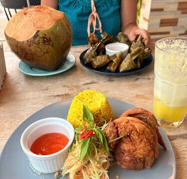 Plate with fried chicken, yellow rice, salad, and sauce; a Siargao-inspired coconut drink and another dish in the background.
