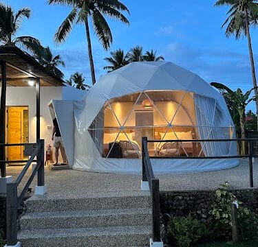 A white geodesic dome with warm lights inside, surrounded by palm trees at dusk in Siargao.