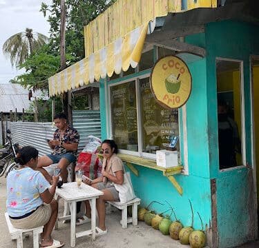 Four people sit at small tables outside a turquoise Siargao café with coconuts lined up on the ground.