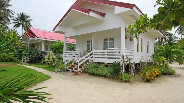 Two white bungalows with red roofs in Siargao, surrounded by tropical plants and palm trees on a sandy path.