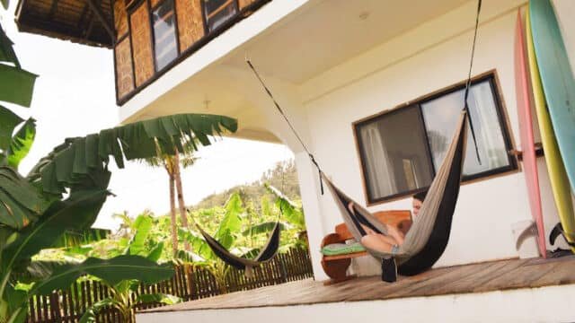 Person relaxing in a hammock on a wooden porch with surfboards nearby, soaking in the laid-back Siargao vibes and lush banana trees.
