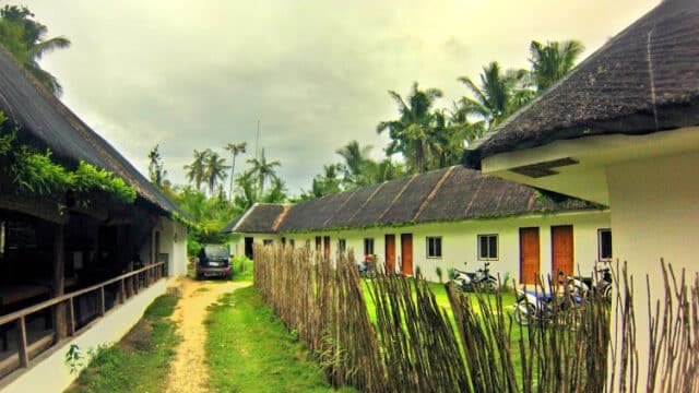 Thatched-roof cottages and palm trees line a dirt path in Siargao; motorbikes are parked near the buildings under a cloudy sky.