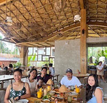 Five people sit around a table with food and drinks in a tropical, open-air Siargao restaurant with a thatched roof.