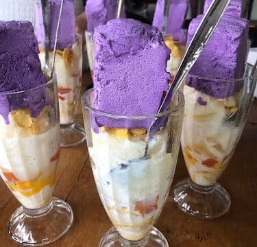 Tall glasses of halo-halo topped with bright purple ube ice cream, Siargao style, spoons inserted, on a wooden table.