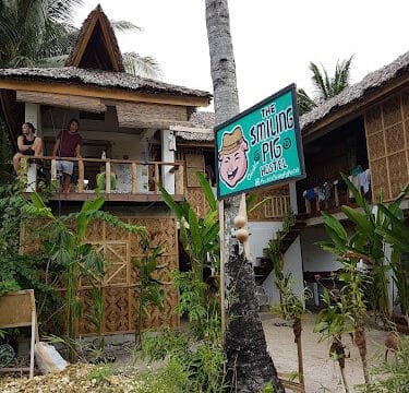 A sign reading The Smiling Pig Hostel is posted on a palm tree in front of bamboo buildings and Siargao greenery.