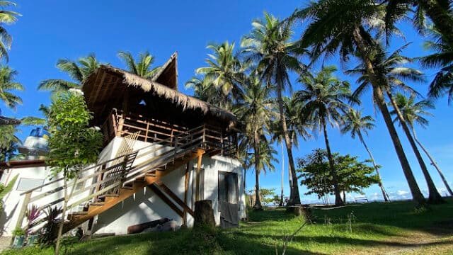 A tropical wooden house with a thatched roof in Siargao, surrounded by palm trees under a bright blue sky.