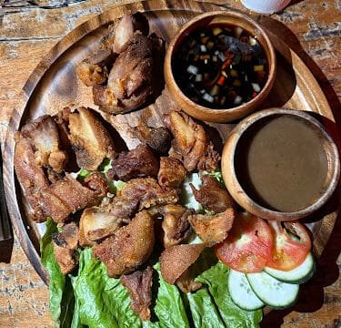 A wooden plate with fried meat, lettuce, cucumber, tomato, and two dipping sauces on a rustic Siargao table.