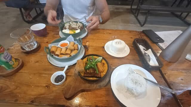 A person at a wooden table in Siargao with spring rolls, rice, soup, and vegetables, ready to eat.