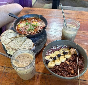 Breakfast on a wooden table in Siargao: shakshuka, bread, smoothie bowls with fruit, and two iced drinks with straws.