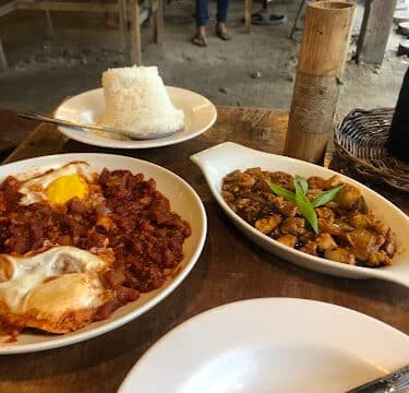 A wooden table in Siargao with rice, fried eggs with meat, a vegetable dish, and an empty plate with a fork.