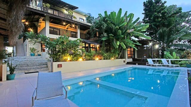 Modern Siargao villa with balconies, tropical plants, and a swimming pool surrounded by lounge chairs at sunset.